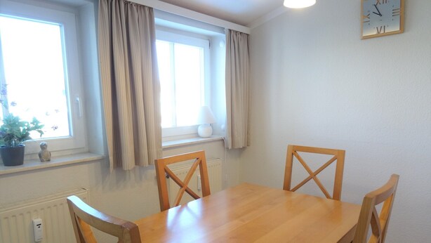 Een eetkamer met een houten tafel en vier stoelen, een raam met gordijnen, een radiator en een wandklok.