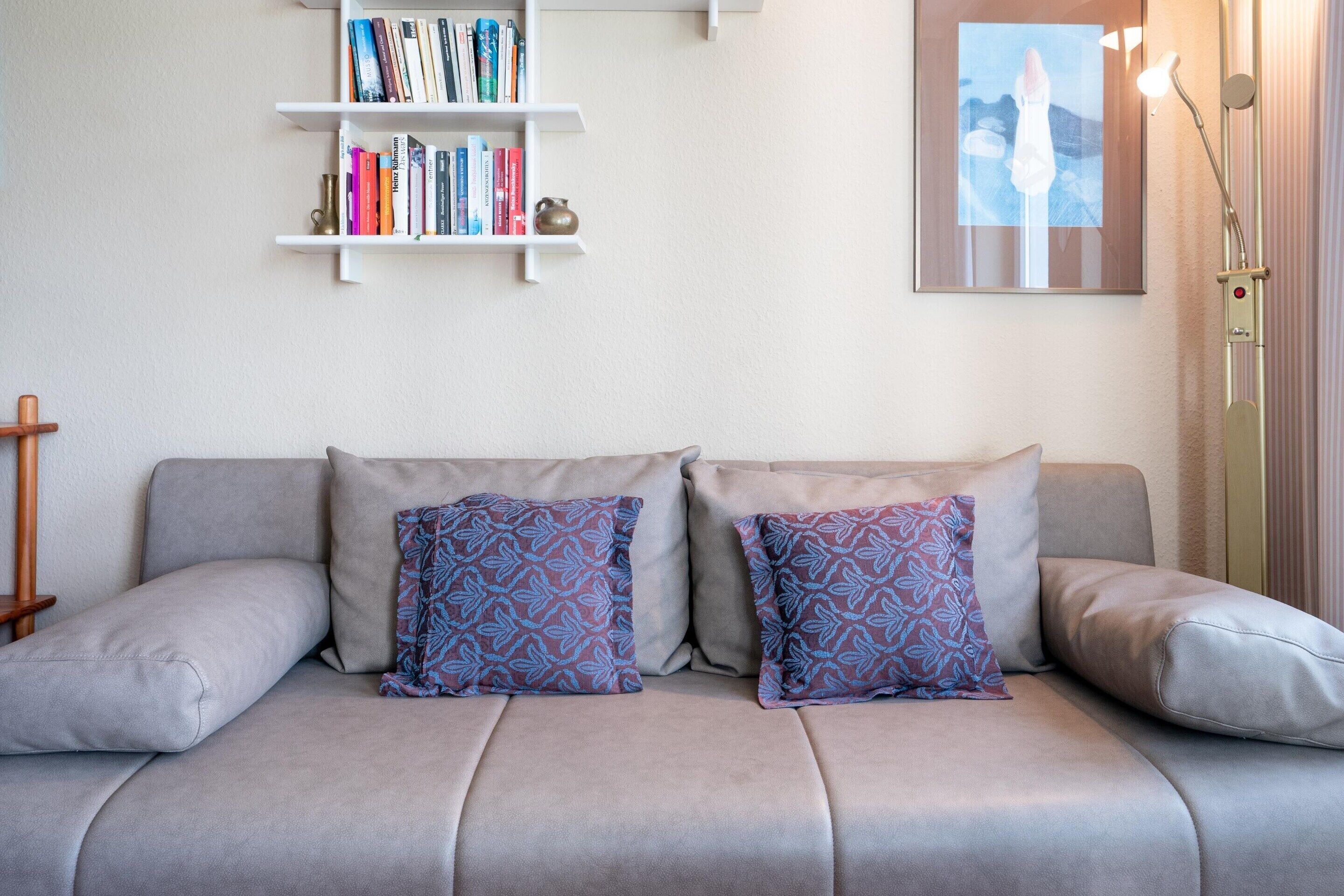 Ein modernes Wohnzimmer mit einer grauen Couch, gemusterten Kissen, einem Bücherregal und einem gerahmten Bild an der Wand.
