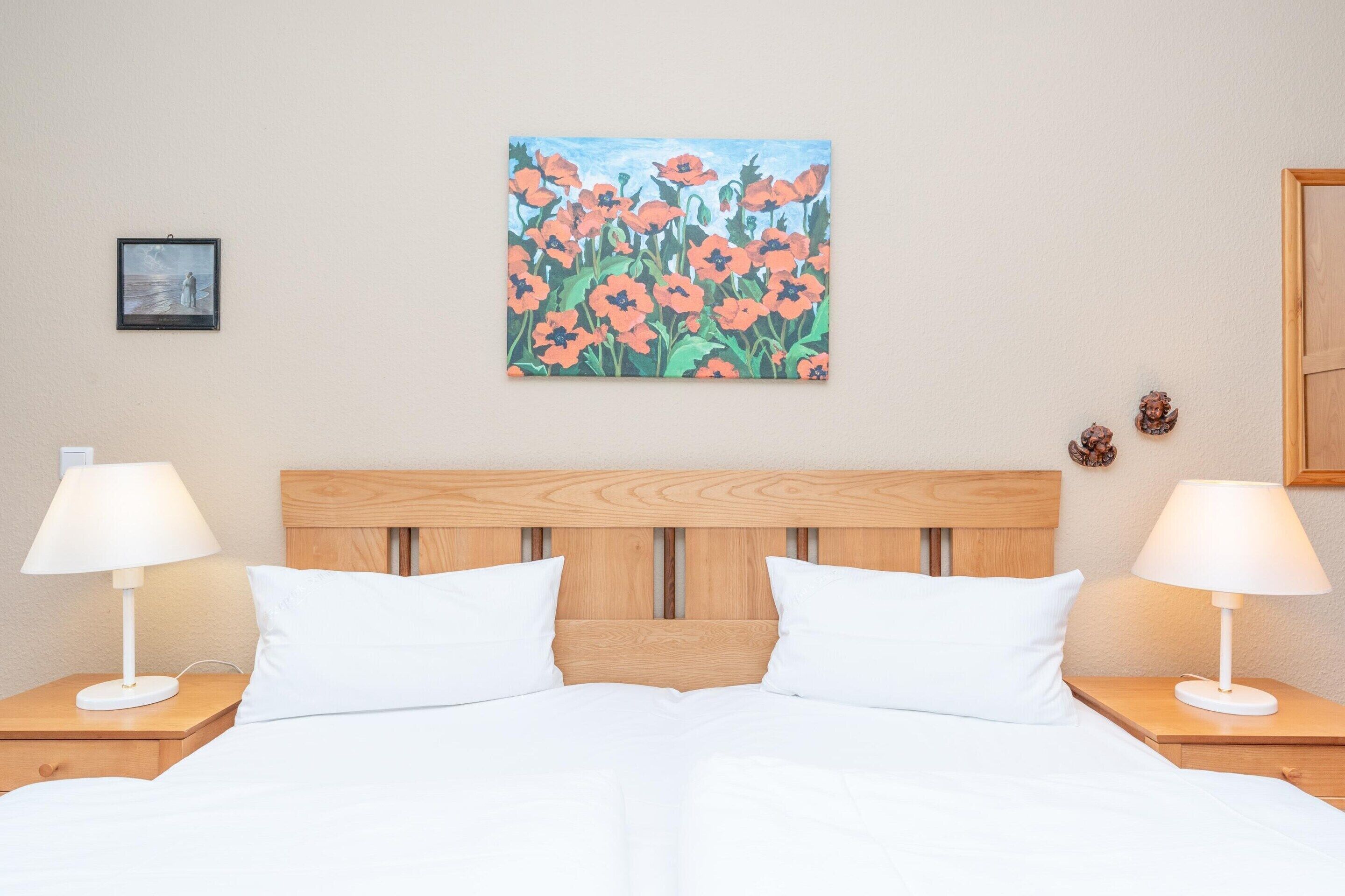 Ein Hotelzimmer mit hölzernem Kopfteil, weißer Bettwäsche, zwei Nachttischlampen und einem Blumenbild an der Wand.
