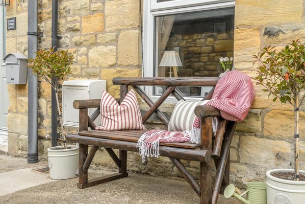 A rustic wooden bench with cushions, potted plants, and a stone building in the background.