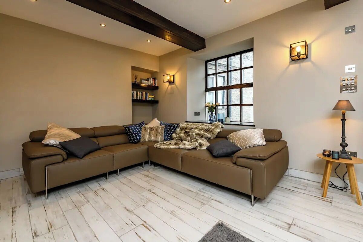 Una sala de estar con un sofá modular marrón, piso de madera, una ventana con persianas, una estantería y una lámpara.