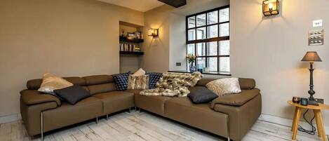 Una sala de estar con un sofá modular marrón, piso de madera, una ventana con persianas, una estantería y una lámpara.