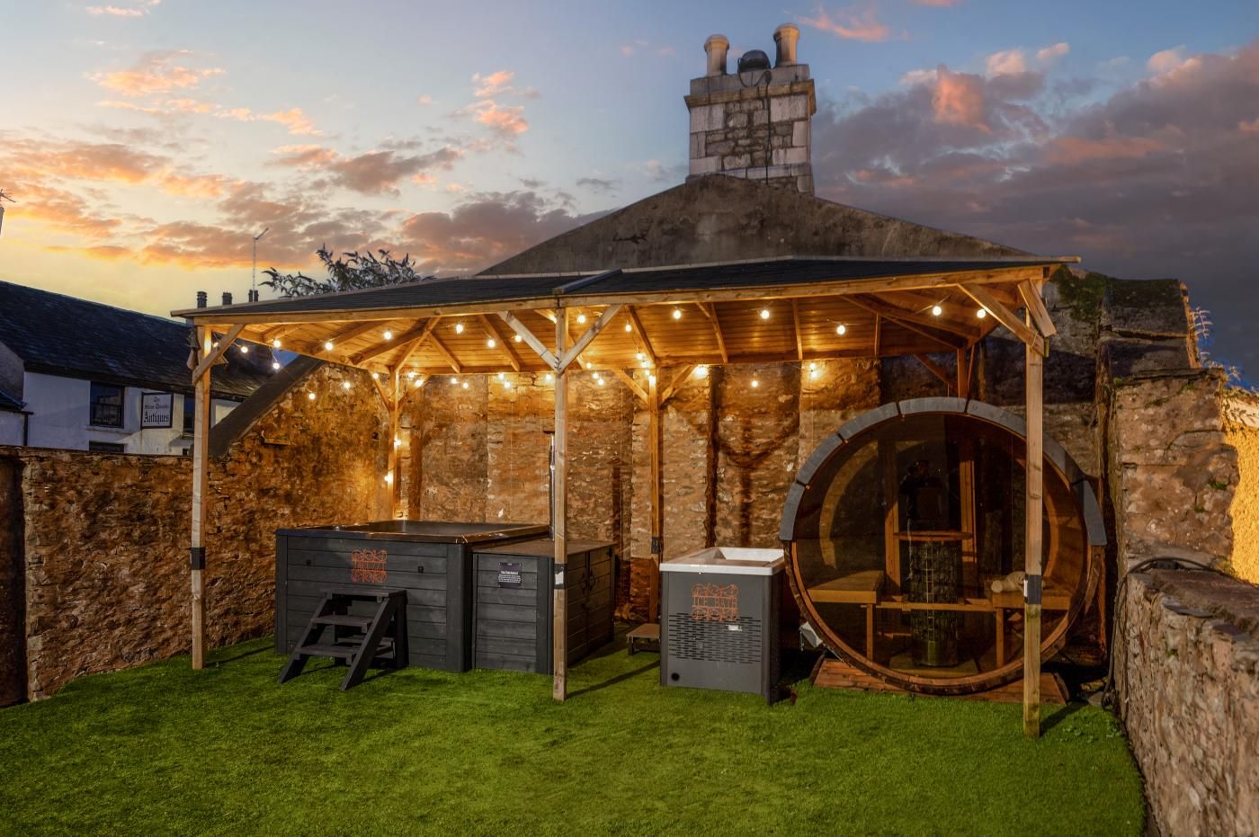 A rustic outdoor seating area with a stone wall backdrop, a wooden pergola with string lights, and a large barrel.