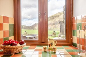 A kitchen with a view of a green field and hills, a wicker basket with fruit, and a pitcher with cups. - 2 Bed in Thwaite (oc-m36624) (Thwaite)