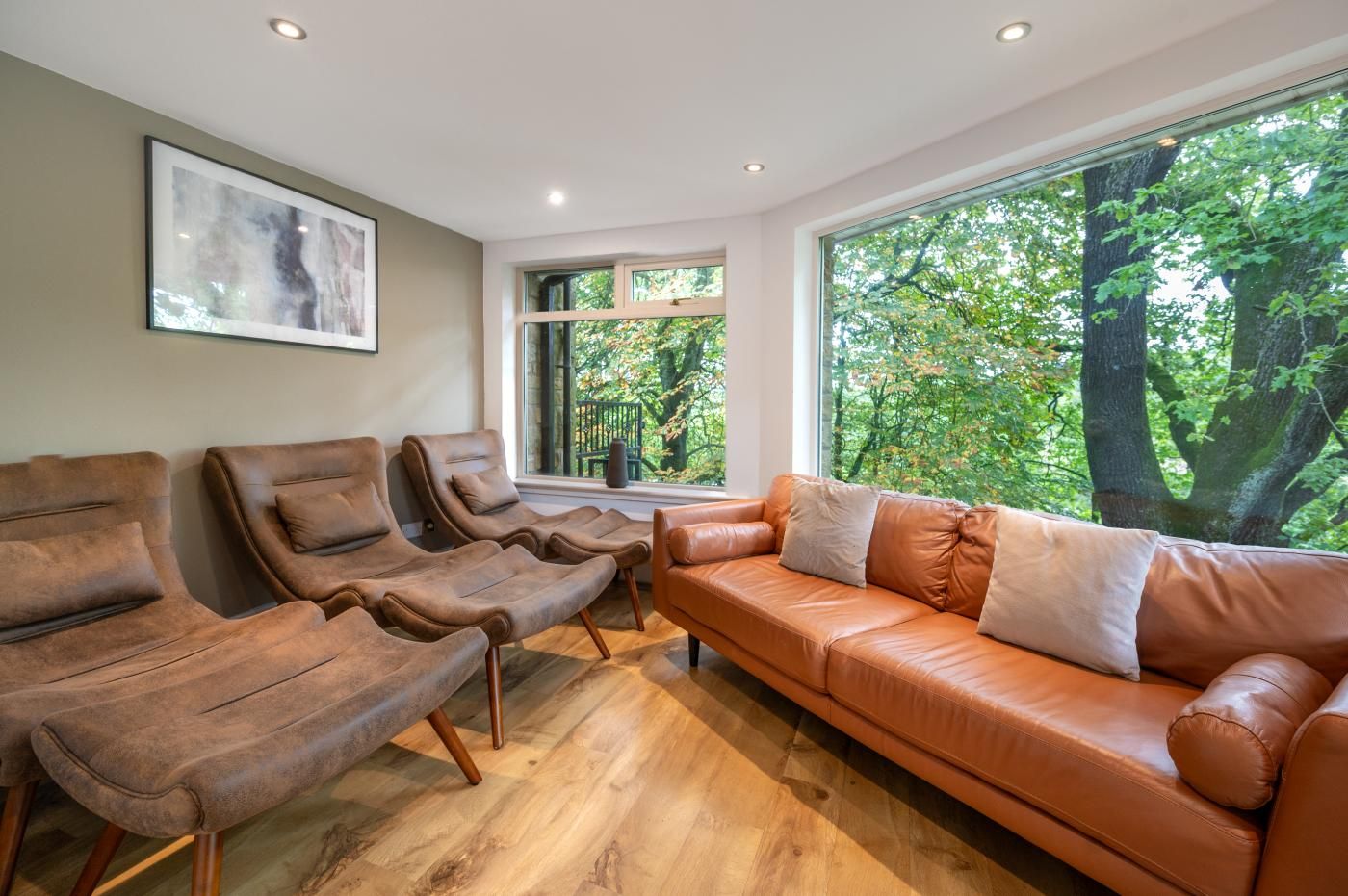 A modern living room with a leather sofa, two recliners, and a large window offering a view of trees.