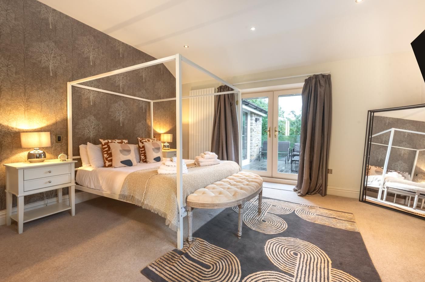 Une chambre à coucher avec un lit à baldaquin, une table de chevet, une commode, un banc, un grand miroir et une vue sur l’extérieur.