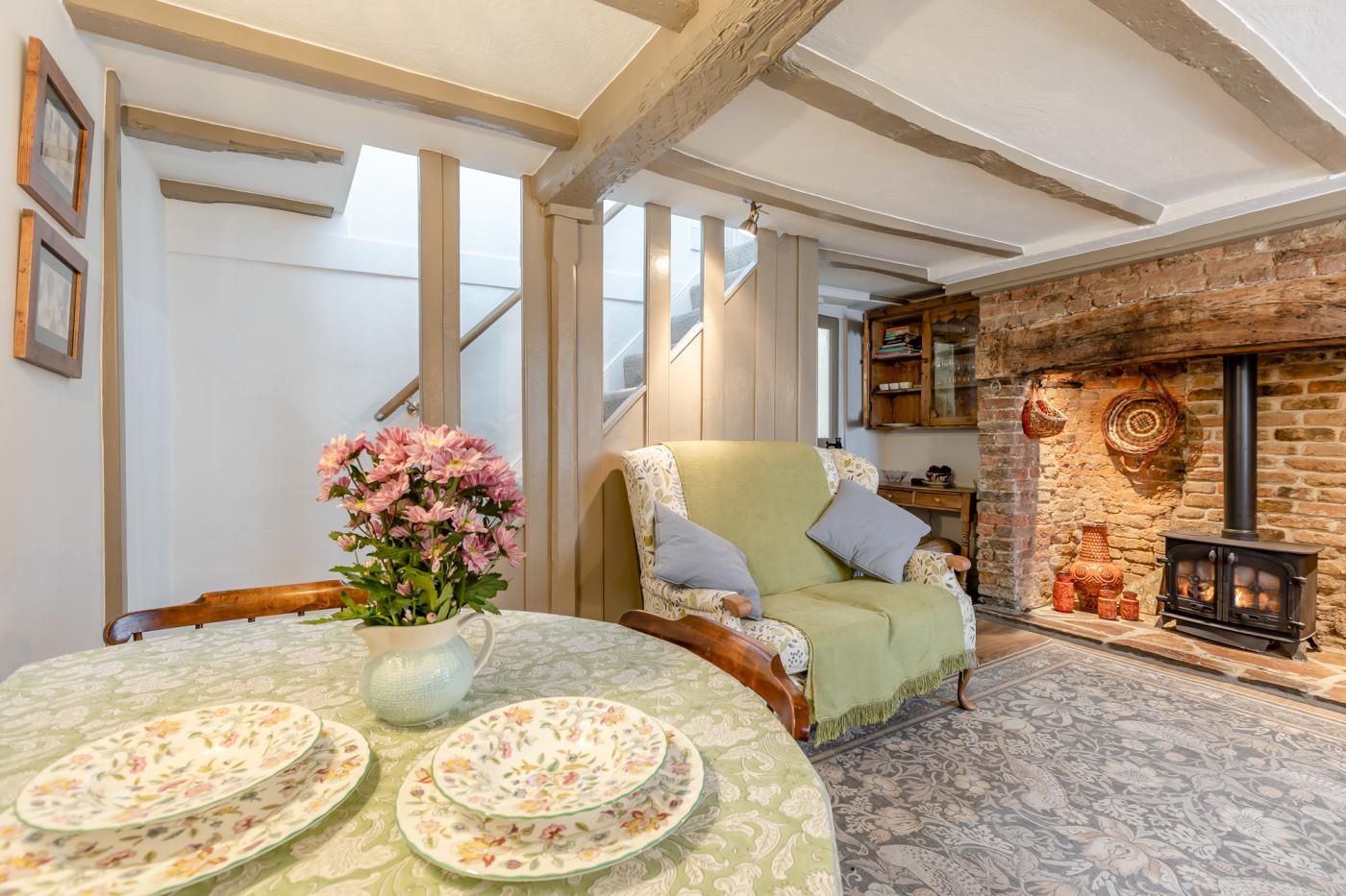 A cozy living room with a fireplace, a dining table with floral tableware, a green armchair, and a staircase leading upstairs.