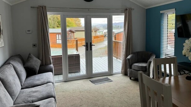 A living room with a grey sofa, a white chair, a TV, and a sliding glass door leading to a deck. - 2 Bed in Troutbeck Bridge (oc-p36885) (Troutbeck Bridge)