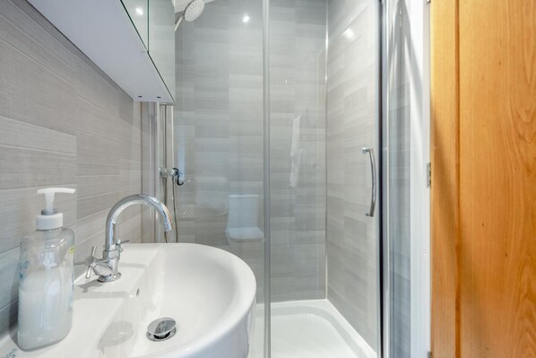 A bathroom with a glass shower enclosure, a white sink with a soap dispenser, and a wooden door.