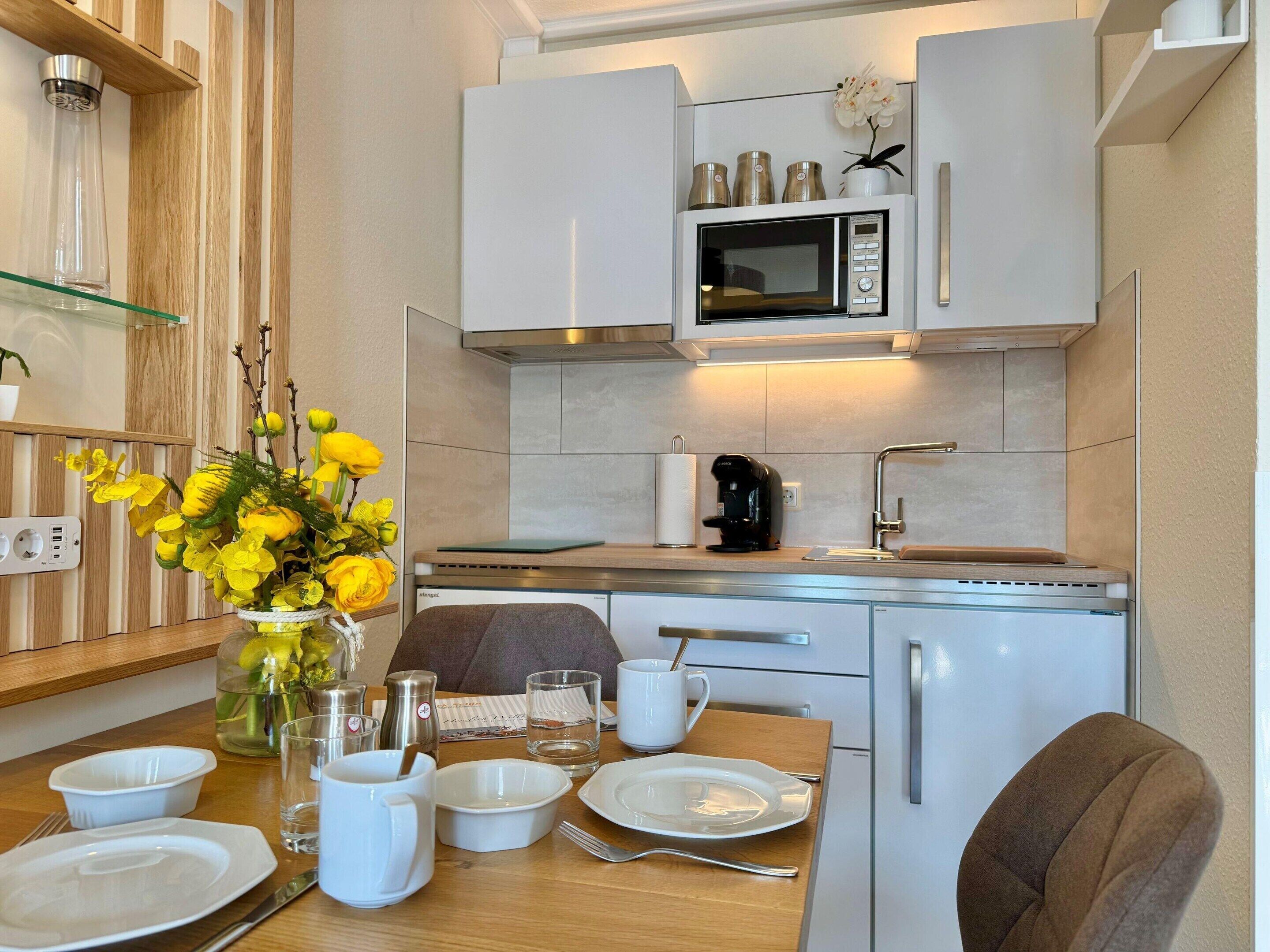 Una cocina moderna con armarios blancos, microondas, cafetera y una mesa de comedor con platos, fuentes y un florero con flores amarillas.