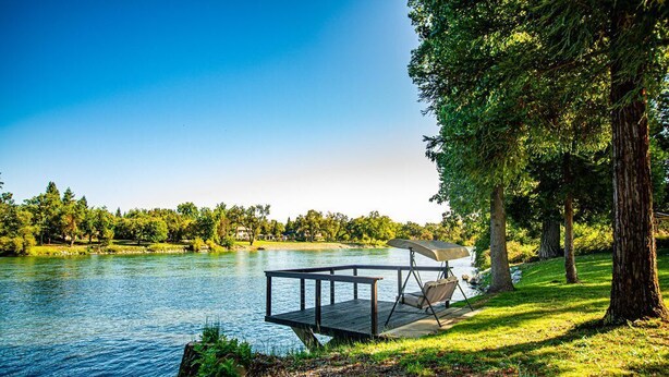 A lakeside deck with a chair and umbrella, surrounded by trees and a clear sky.