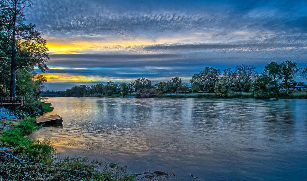 Ein Fluss mit einer hölzernen Anlegestelle, Bäumen und einem bewölkten Himmel.
