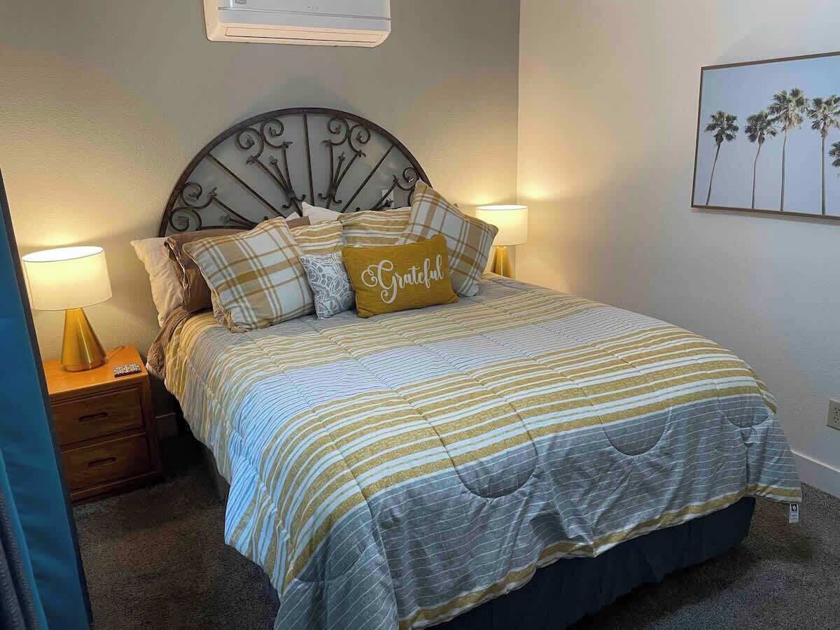 Ein Bett mit Metall-Kopfteil, gestreifter Bettwäsche und einem Kopfkissen mit der Aufschrift 'Grateful'.