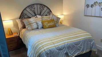 Ein Bett mit Metall-Kopfteil, gestreifter Bettwäsche und einem Kopfkissen mit der Aufschrift 'Grateful'.