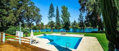 A rectangular swimming pool with a wooden deck and lounge chairs, surrounded by trees and a lake view.
