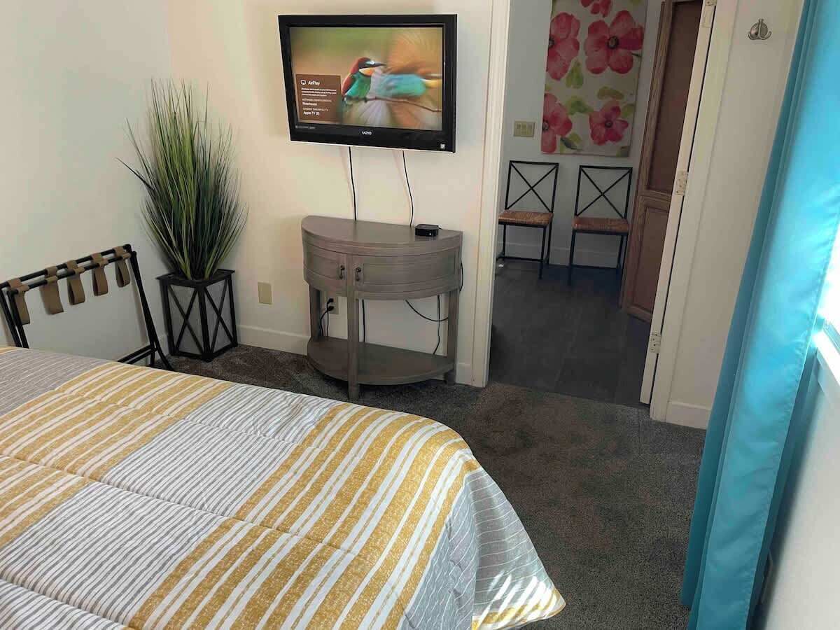 A bedroom with a bed, a TV on the wall, a small table, a chair, and a potted plant.