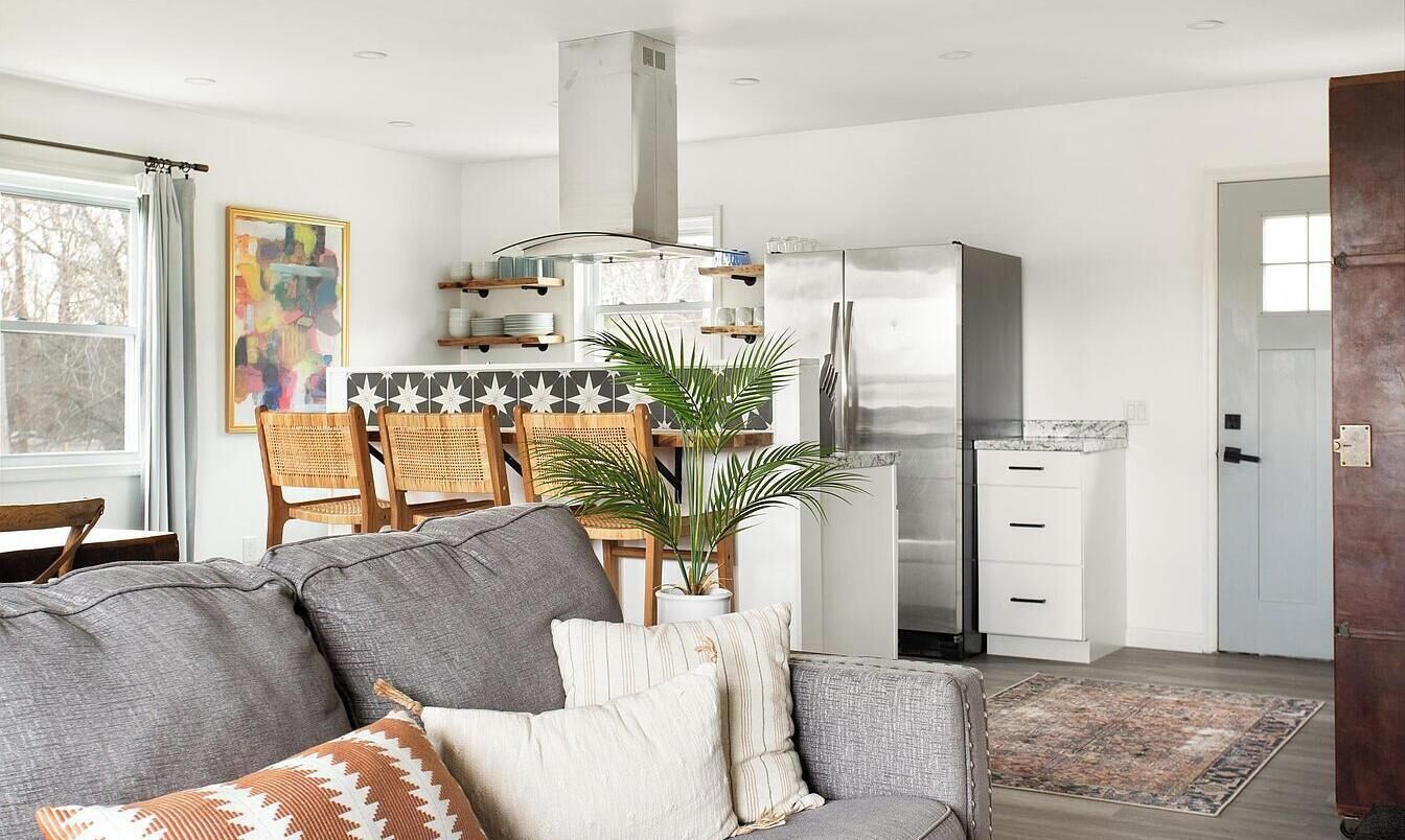 Et moderne køkken med et rustfrit stål køleskab, et spiseområde med et bord og stole, og en stue med en sofa og dekorative puder.