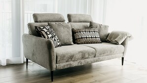 Um sofá cinzento com almofadas estampadas em preto e branco, posicionado num quarto com soalho de madeira e uma janela grande com cortinas leves.