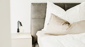 Cama bem arrumada com roupa de cama branca e duas almofadas, abajur e mesa de cabeceira branca.