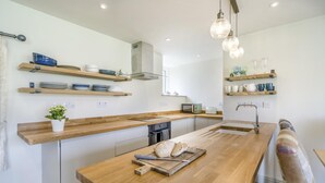 Een moderne keuken met houten aanrechtbladen, een centraal eiland, hangende lampen en inbouwplanken met servies en bekers.