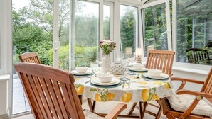 Un comedor en una estructura de cristal con una mesa dispuesta para cuatro personas, rodeada de sillas de madera y grandes ventanales que ofrecen vistas a la vegetación.
