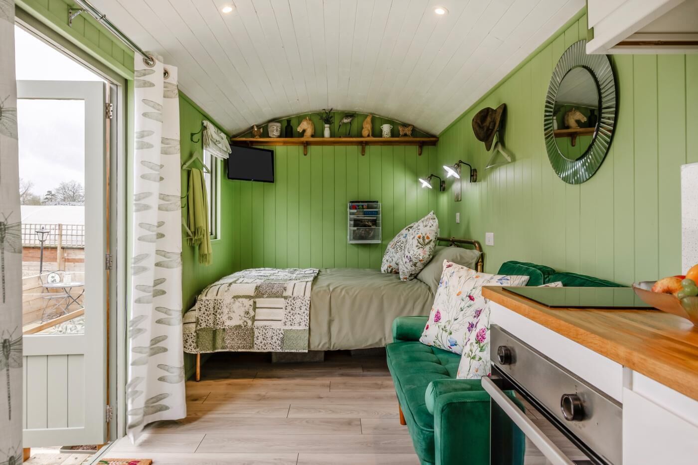 Quarto com cama, sofá verde, mesa de madeira, espelho e janela com cortinas.