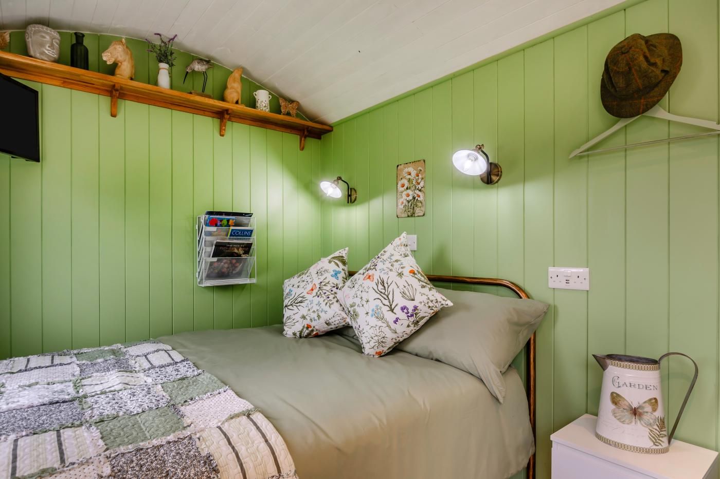 A green-themed bedroom with a bed, pillows, a shelf with decorative items, a lamp, and a wall-mounted light fixture.