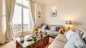A living room with a glass coffee table, beige sofas, a view of the sea through large windows, and a radiator. - 3 Bed in Ilfracombe (oc-h36896) (Ilfracombe)