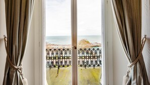 A view through a door with curtains tied back, revealing a balcony with a metal railing and a beach in the background. - 3 Bed in Ilfracombe (oc-h36896) (Ilfracombe)