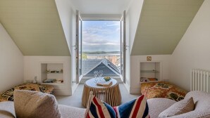 Ein gemütliches Zimmer mit hoher Decke, einem kleinen runden Tisch mit Gläsern, einer Couch mit gemusterten Kissen und Blick auf Gebäude durch ein großes Fenster.