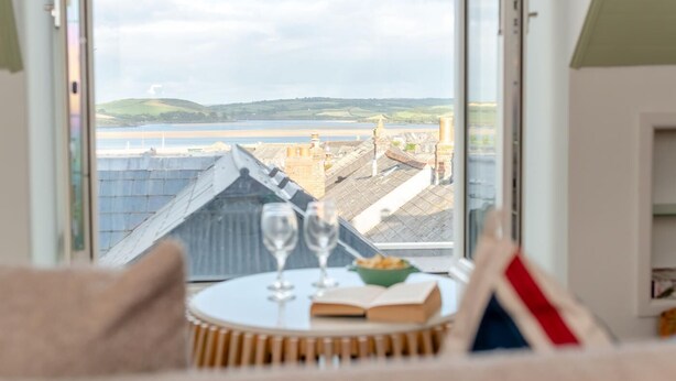 Ein Zimmer mit großem Fenster und Blick auf eine Küstenlandschaft, ein Tisch mit Obstschale und ein Buch.