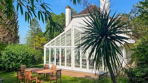 A conservatory with a glass roof, surrounded by lush greenery and a wooden table with chairs. - 4 Bed in Beckley (oc-s34808) (Beckley)