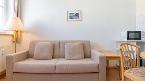 A neutral-toned sofa with two cushions, a wooden chair, a small table, a wall-mounted picture, and a white microwave on a countertop. - Attraktive Ferienwohnung fur Urlaub zu Zweit in Sellin (Sellin)