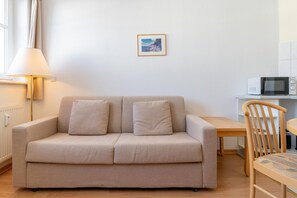 A neutral-toned sofa with two cushions, a wooden chair, a small table, a wall-mounted picture, and a white microwave on a countertop. - Attraktive Ferienwohnung fur Urlaub zu Zweit in Sellin (Sellin)