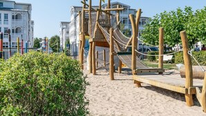 A playground with a wooden slide and climbing structures, surrounded by sand and greenery, with buildings in the background. - Attraktive Ferienwohnung fur Urlaub zu Zweit in Sellin (Sellin)