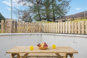 Ein Holztisch mit Picknick-Aufsatz, mit einer Obstschale und zwei Gläsern Orangensaft, vor einer weißen Wand mit einem Holzzaun im Hintergrund.