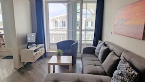 A living room with a sofa, a chair, a small table with a plant, a TV on a stand, and a balcony with a view of buildings. - Geschmackvoll Eingerichtete Ferienwohnung mit Balkon in Sellin (Sellin)