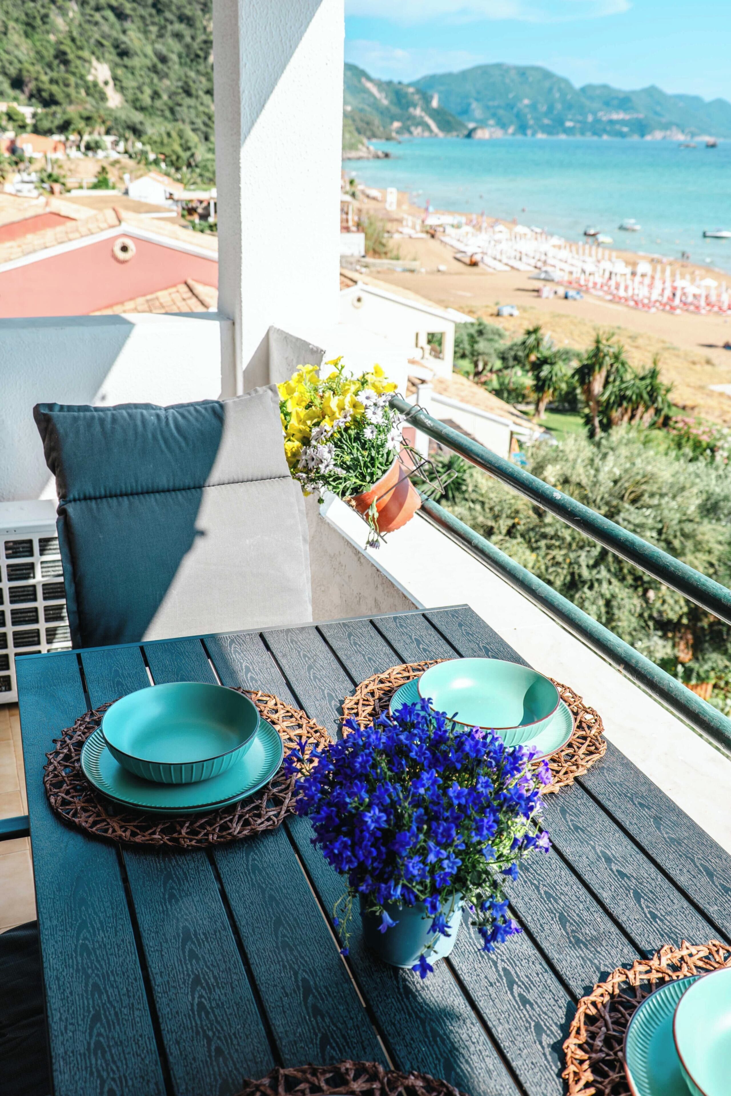 Varanda com mesa posta para dois, planta em vaso e vista para a praia e montanhas.