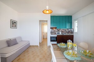 Una cocina moderna con gabinetes color turquesa, una encimera blanca y un microondas. En primer plano, una mesa de comedor con cristalería transparente y un sofá de color claro.