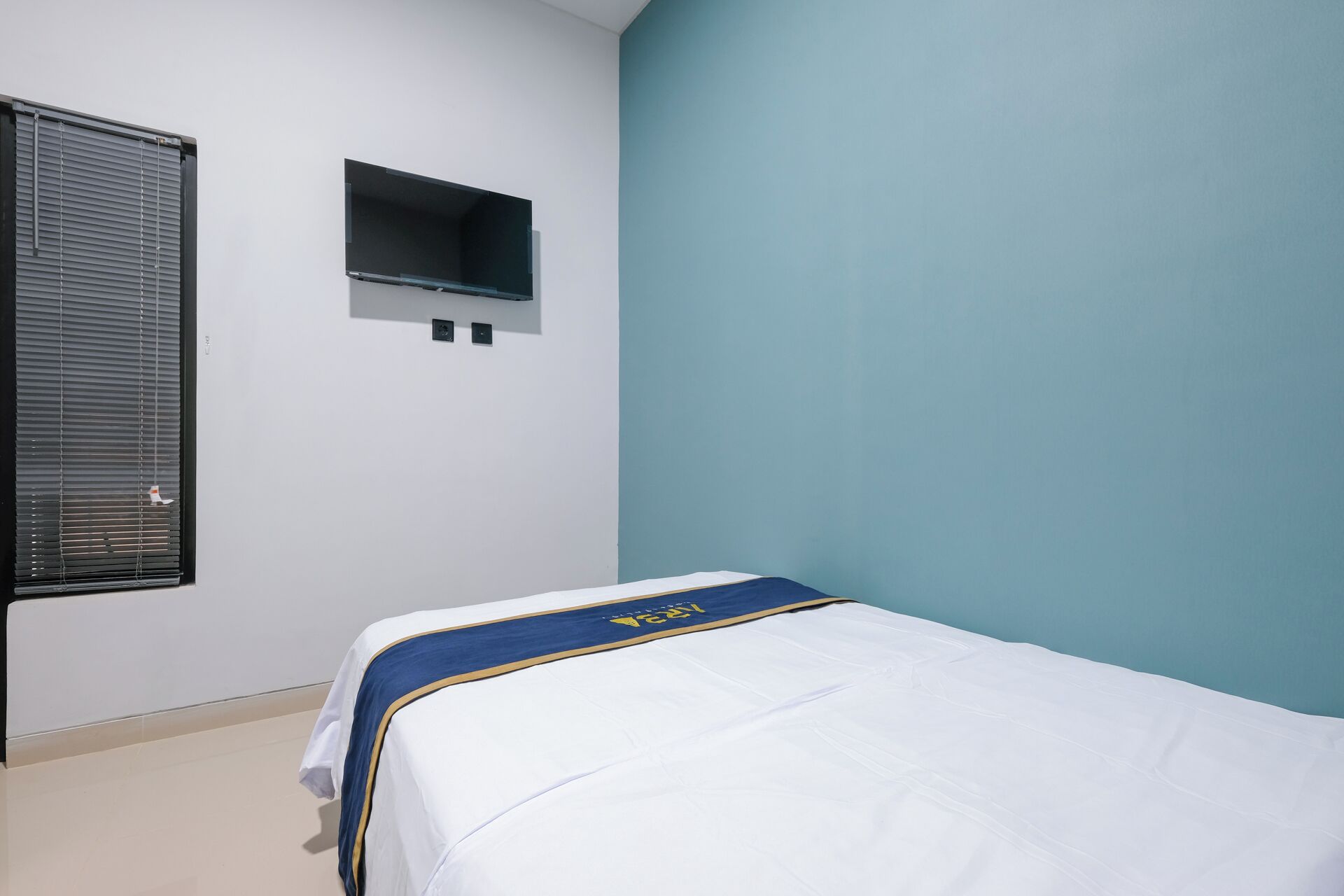 Ein Einzelbett mit weißer Matratze und einer Bettdecke in Blau und Gold, ein kleines Regal und ein Fenster mit Jalousien.