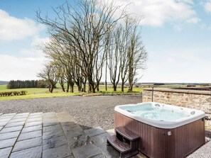 A hot tub with a wooden bench, surrounded by a stone wall and a gravel path, with a view of a field and trees in the background. - 7 Bed in Ellon (oc-ca249) (Methlick)