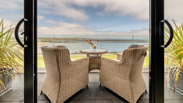 Ein Balkon mit Korbmöbeln, einem Glastisch und Blick auf das Meer und einen Steg.