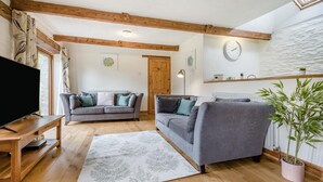 A living room with two grey sofas, a wooden TV stand, a large window with curtains, a clock on the wall, and a potted plant. - 2 Bed in St. Ervan (oc-s36909) (Saint Ervan)