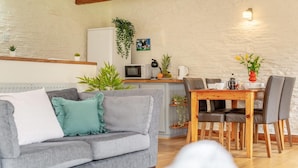 A cozy living room with a grey sofa, a wooden dining table with chairs, a white refrigerator, a microwave, and a small kitchen area with plants and a coffee maker. - 2 Bed in St. Ervan (oc-s36909) (Saint Ervan)