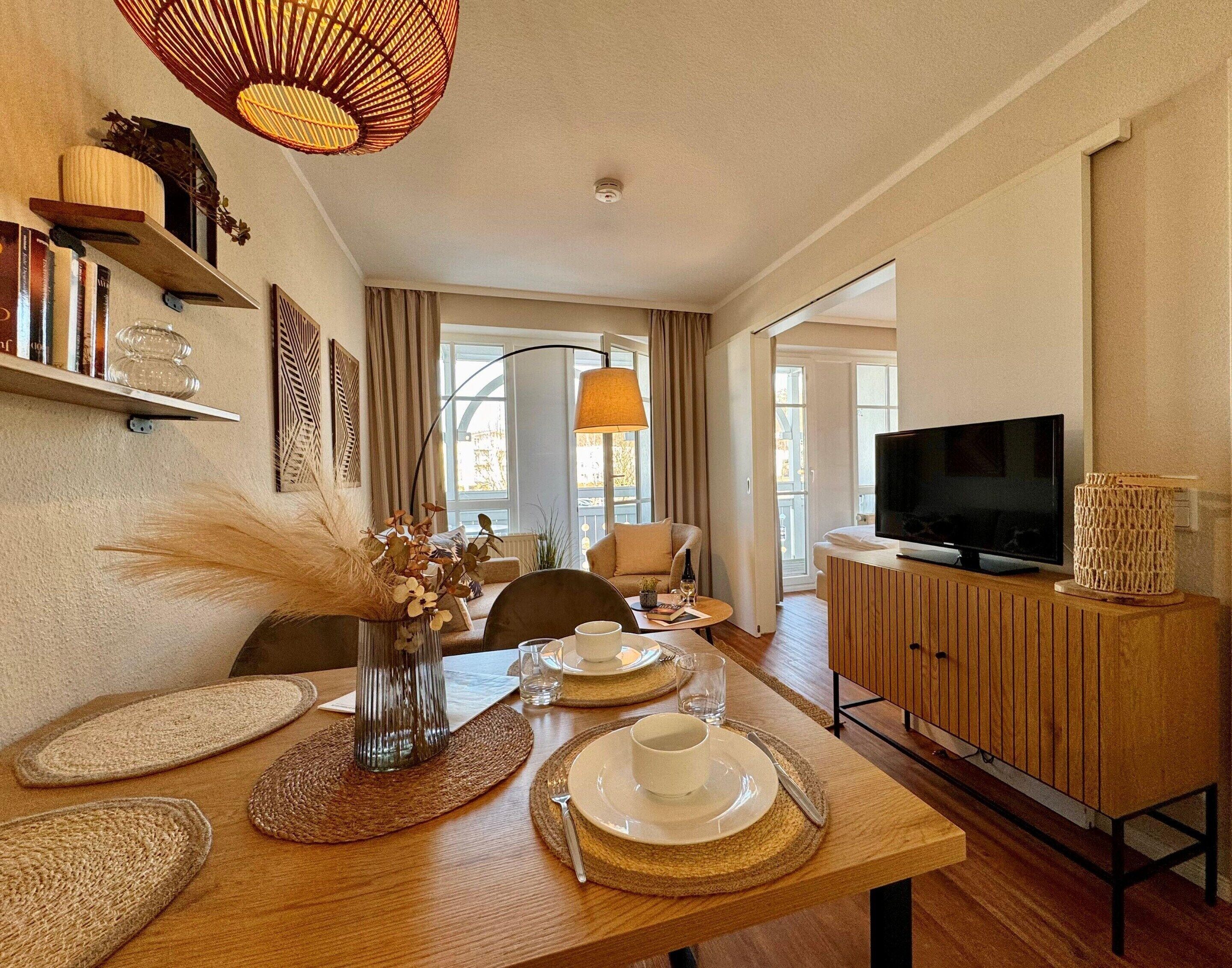 A modern living room with a dining table set, a television on a wooden cabinet, and a large window with curtains.