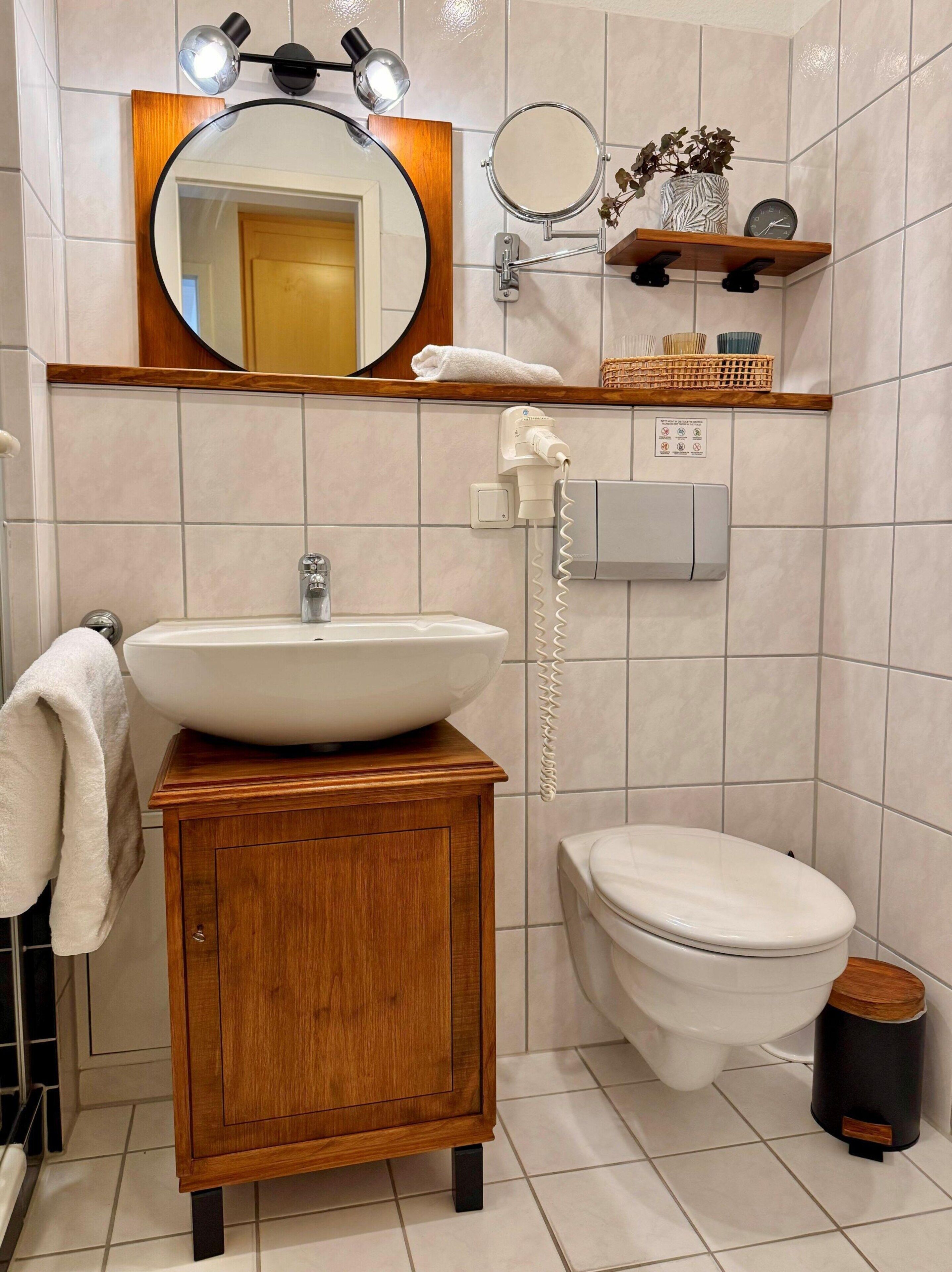 A bathroom with a wooden vanity, a large round mirror, a sink, a toilet, a towel rack, a shelf with a plant, and a waste bin.