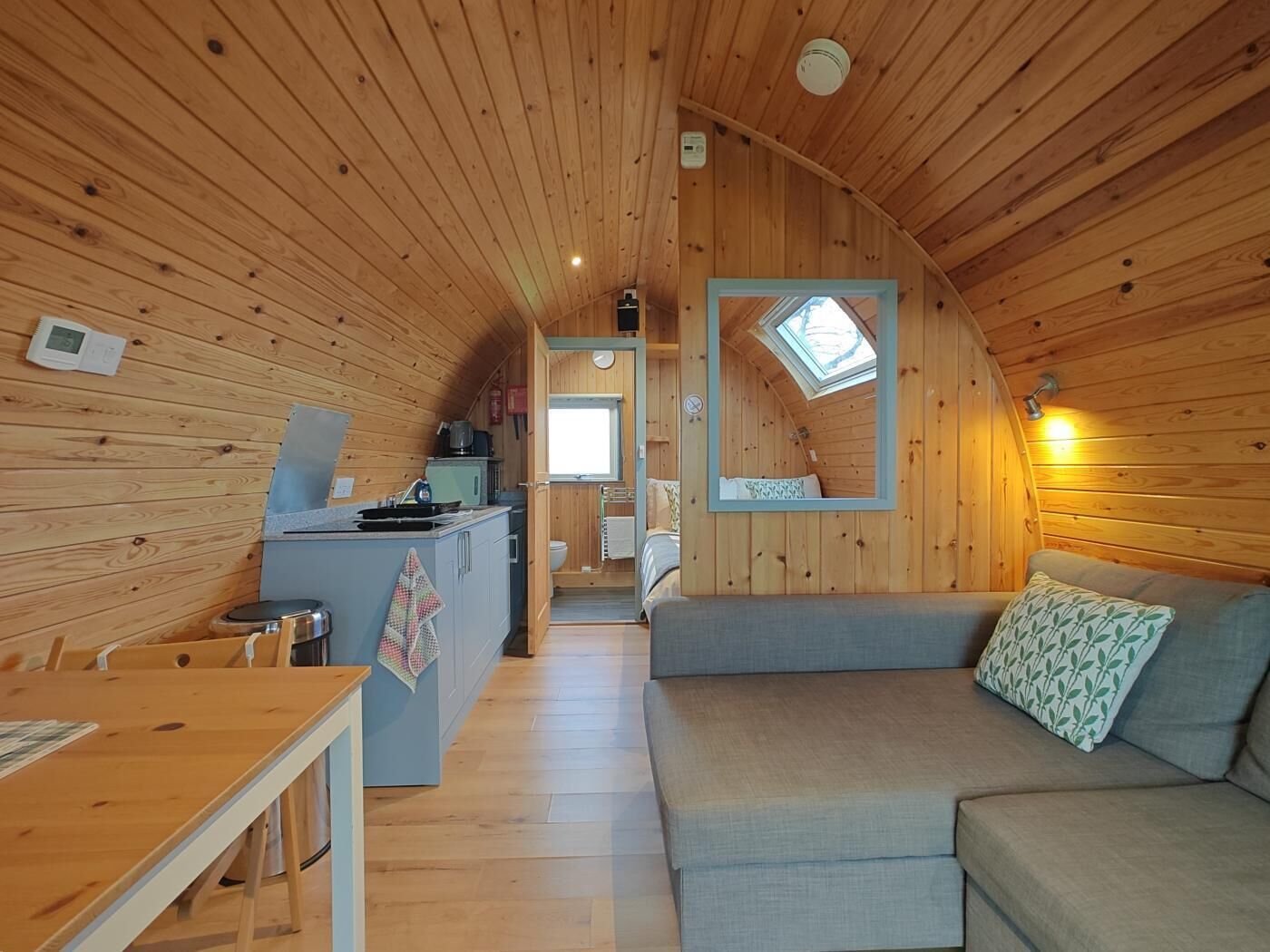 L’intérieur chaleureux d’un chalet, avec un revêtement mural en bois, une petite cuisine, un coin repas et un salon comprenant un canapé et une fenêtre.