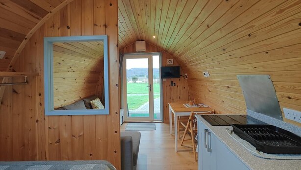 Interior acogedor de una cabaña con techo de madera, una cama, una pequeña cocina y vistas al exterior.