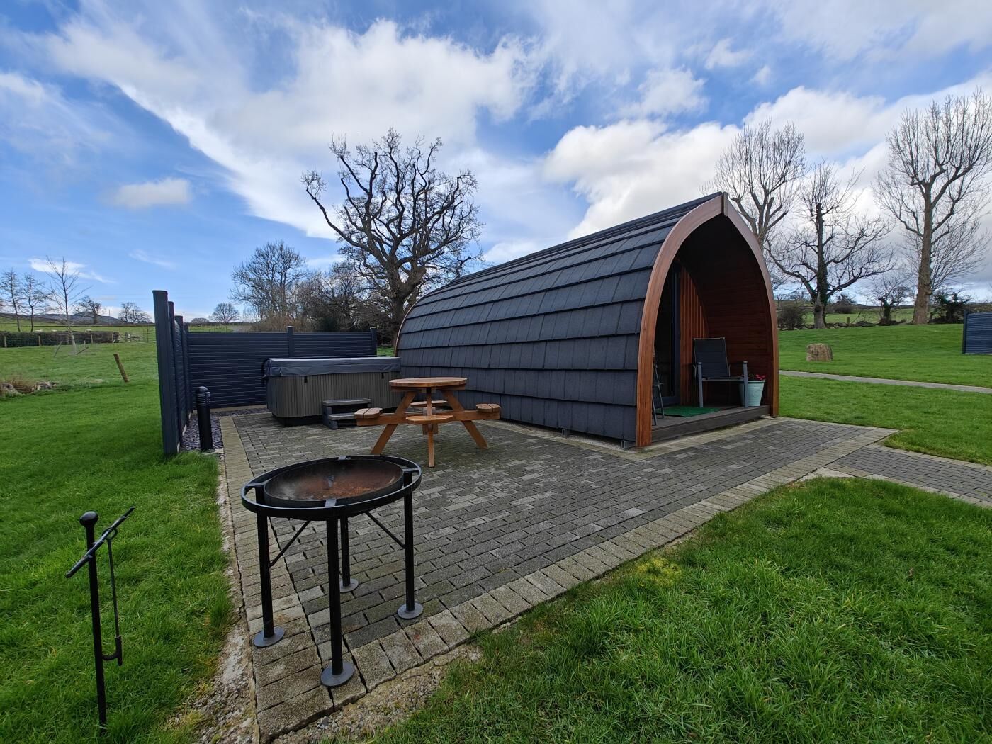 Un espace extérieur rustique comprenant une table et des bancs en bois, un jacuzzi et une structure couverte dotée d’un toit incurvé.