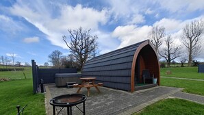 Un espacio exterior rústico con una mesa de madera y bancos, una bañera de hidromasaje y una estructura cubierta con techo curvo.